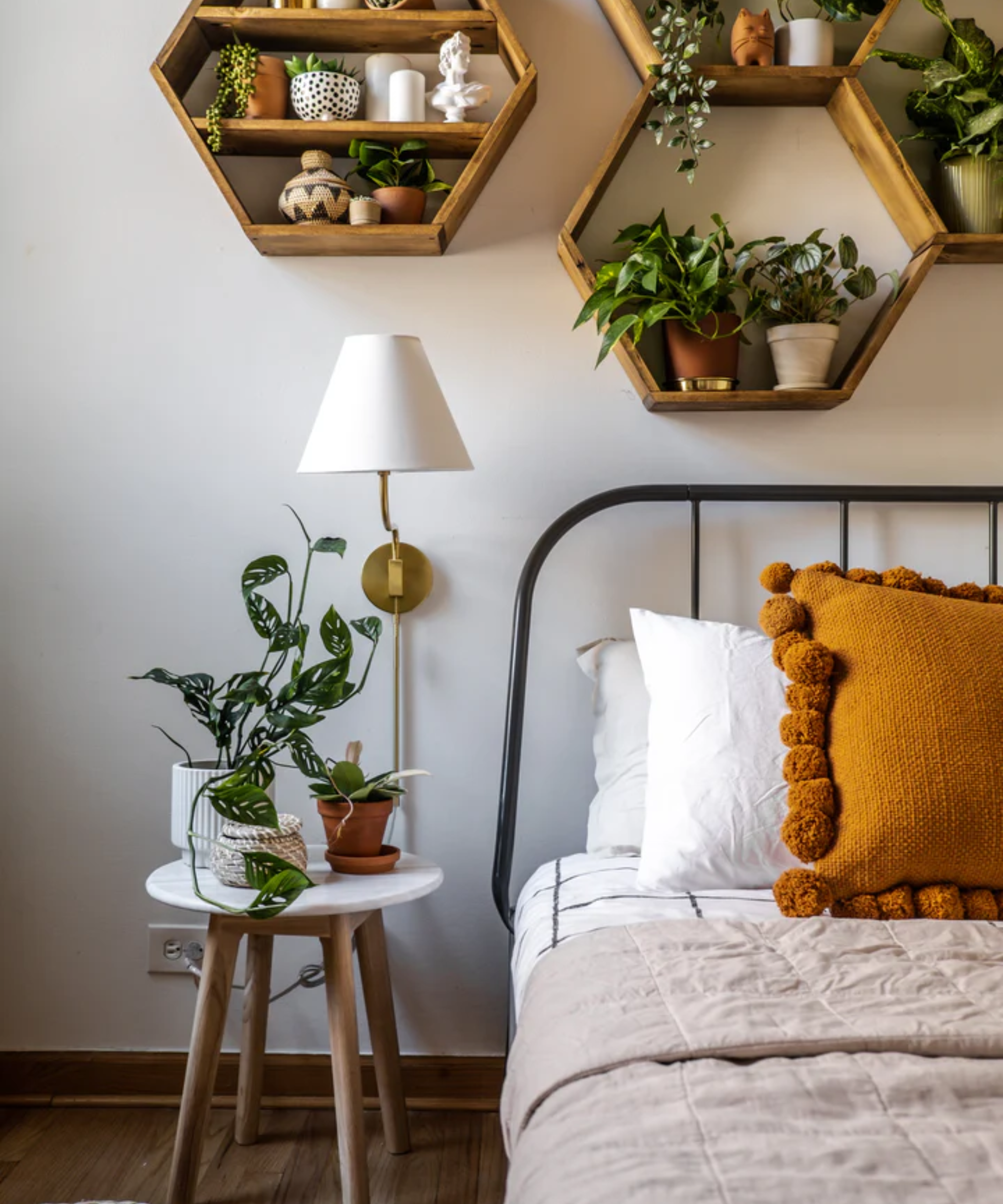 a cozy bedroom with lots of plants, a metal bedframe, a small side table, and a plug in sconce on the wall