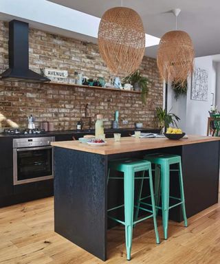 A kitchen with a black kitchen island with a wooden counter, two seagrass lights above it and two aqua stools underneath it, and black cabinets and a brick wall to the back