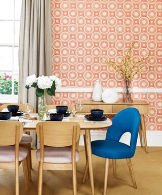 A dining room with peach and white wallpaper, a window with long gray curtains, a wooden dining table with navy blue dinnerware and wooden chairs underneath it, and a wooden sideboard and bookcase beyond it