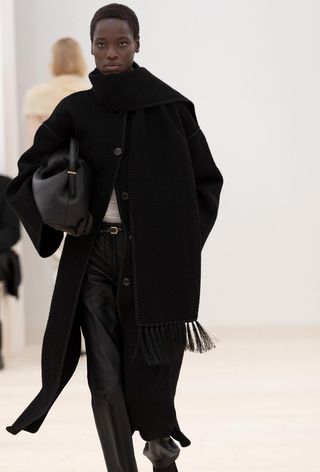 One of the best jacket coat brands, Toteme, is shown in an image of a model wearing a black scarf coat with leather trousers and a clutch bag