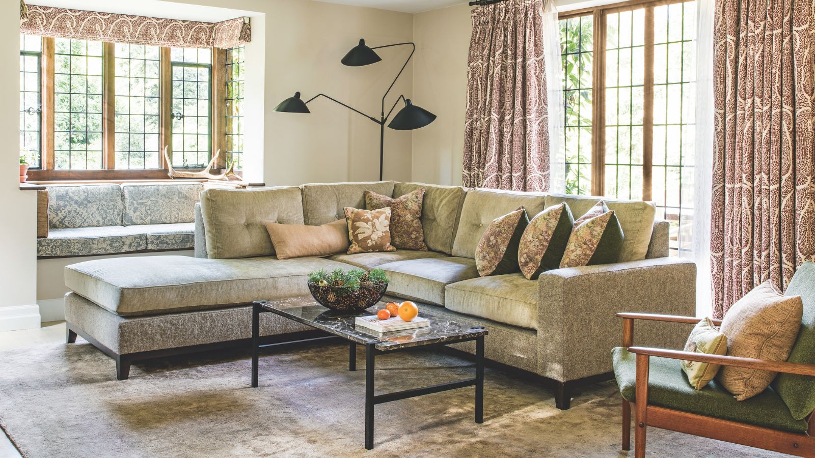 A cozy living room with a large L-shaped velvet couch with cushions, a large velvet rug, marble coffee table, and large windows with patterned curtains. In the back corner is a large black lamp, and in the foreground is a green chair with a wooden frame and two cushions.