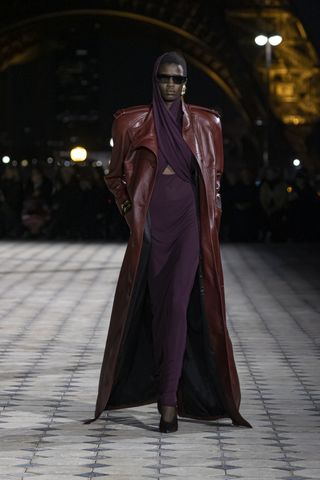 A female model wearing a brown leather coat and a long purple dress walking down a runway.