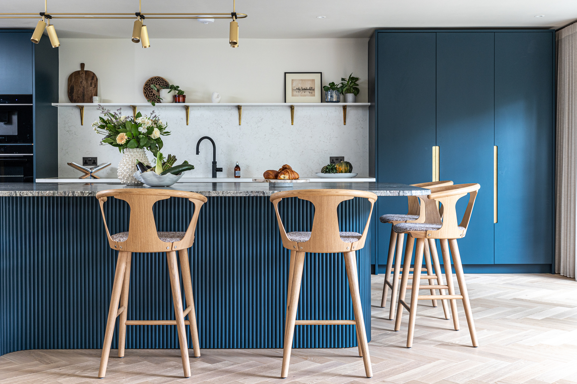 modern kitchen with blue joinery, white marble backsplash and countertop, timber bar stools around the island, brass lights overhead, and an open shelf above the stove