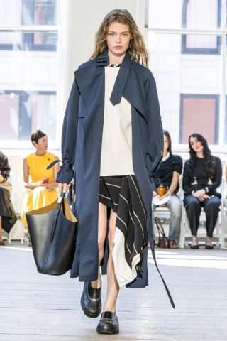 A photo of a model walking for Proenza Schouler wearing black clogs and a sapphire blue trench coat