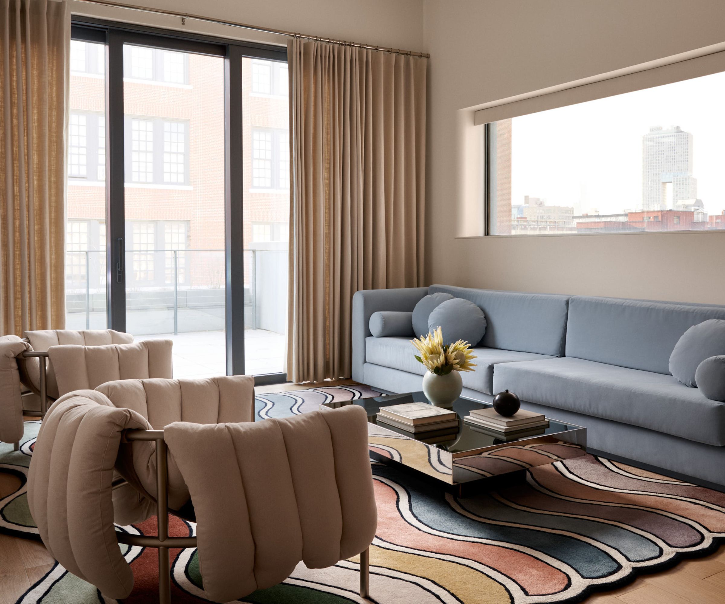 Modern living room with glass doors overlooking NYC skyline, blue long sofa with round cushions, colorful abstract rug, glass coffee table with decor and quilted modern armchairs