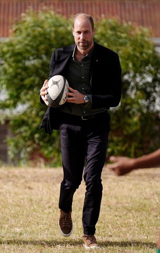 Prince William wearing a blue blazer and pants running with a rugby ball outside