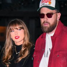 Taylor Swift has bright red lipstick and brown hair as she walks with Travis Kelce who is wearing a baseball cap