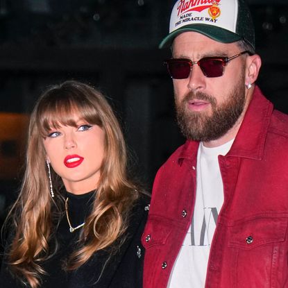 Taylor Swift has bright red lipstick and brown hair as she walks with Travis Kelce who is wearing a baseball cap