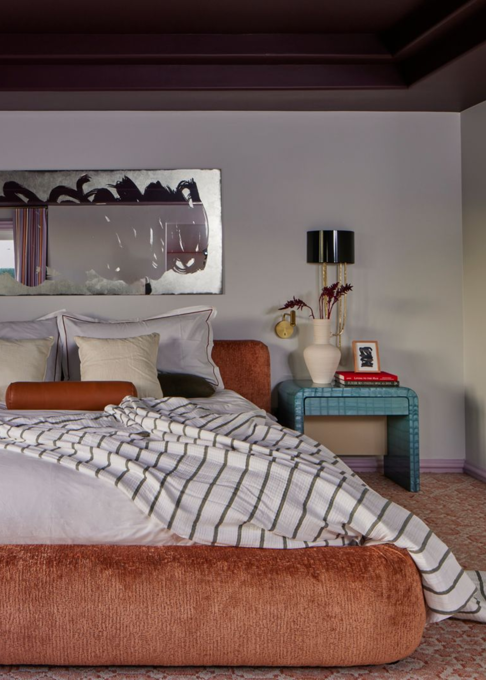 A white bedroom with a chrome mirror, a tiled blue side table, a vase with stems, a stack of books, a wall lamp, and a bed with a striped throw, a set of rectangular pillows and a tan bolster pillow