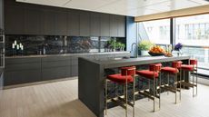 Kitchen with pale wood flooring, dark grey kitchen cupboards, kitchen island with black and white marbled worktop and splash backs. Metal bar stool with upholstered orange seats, floor to ceiling windows onto the balcony. 