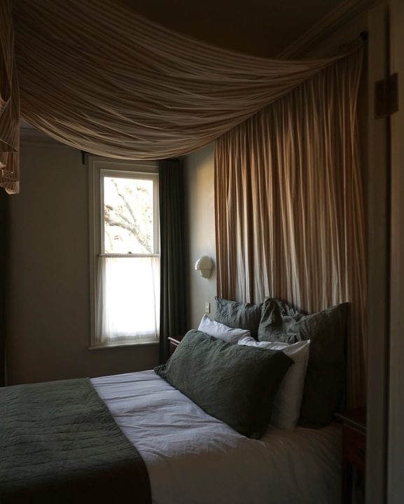 Image of a dark bedroom that has olive green and white bedding. There is a canopy/drapery hanging over the bed that is an orange-beige color.