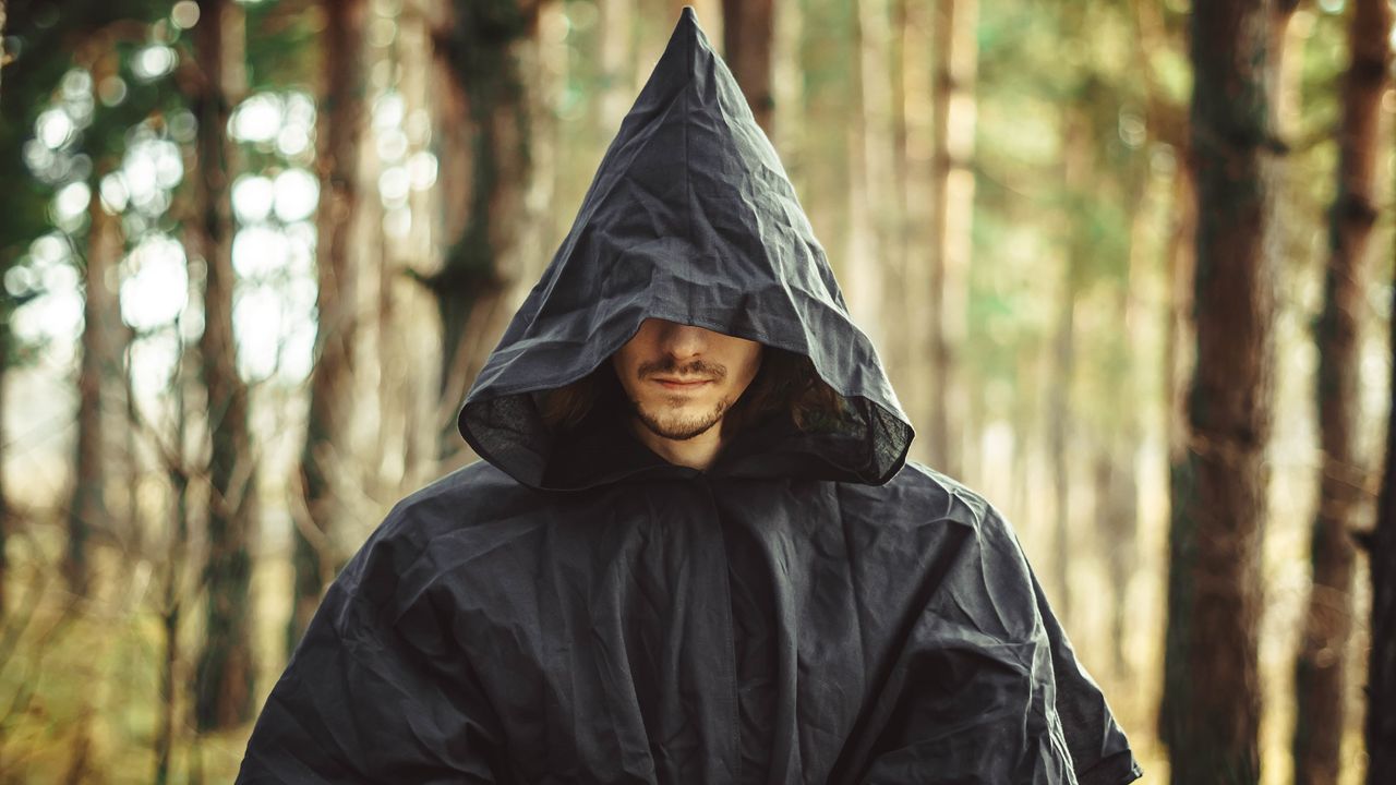 A somewhat sinister-looking man wears a hooded black cloak.