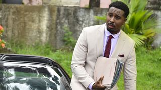 Sterling Fox (Trieve Blackwood-Cambridge), wearing a cream-coloured suit with a light pink shirt and a red tie, is leaning against his open-top sports car and carrying several police case files in his right hand