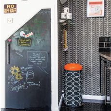 corner of the kitchen with black tiled floor, Orla Kiely patterned wallpaper and bin and cupboard door painted with blackboard paint