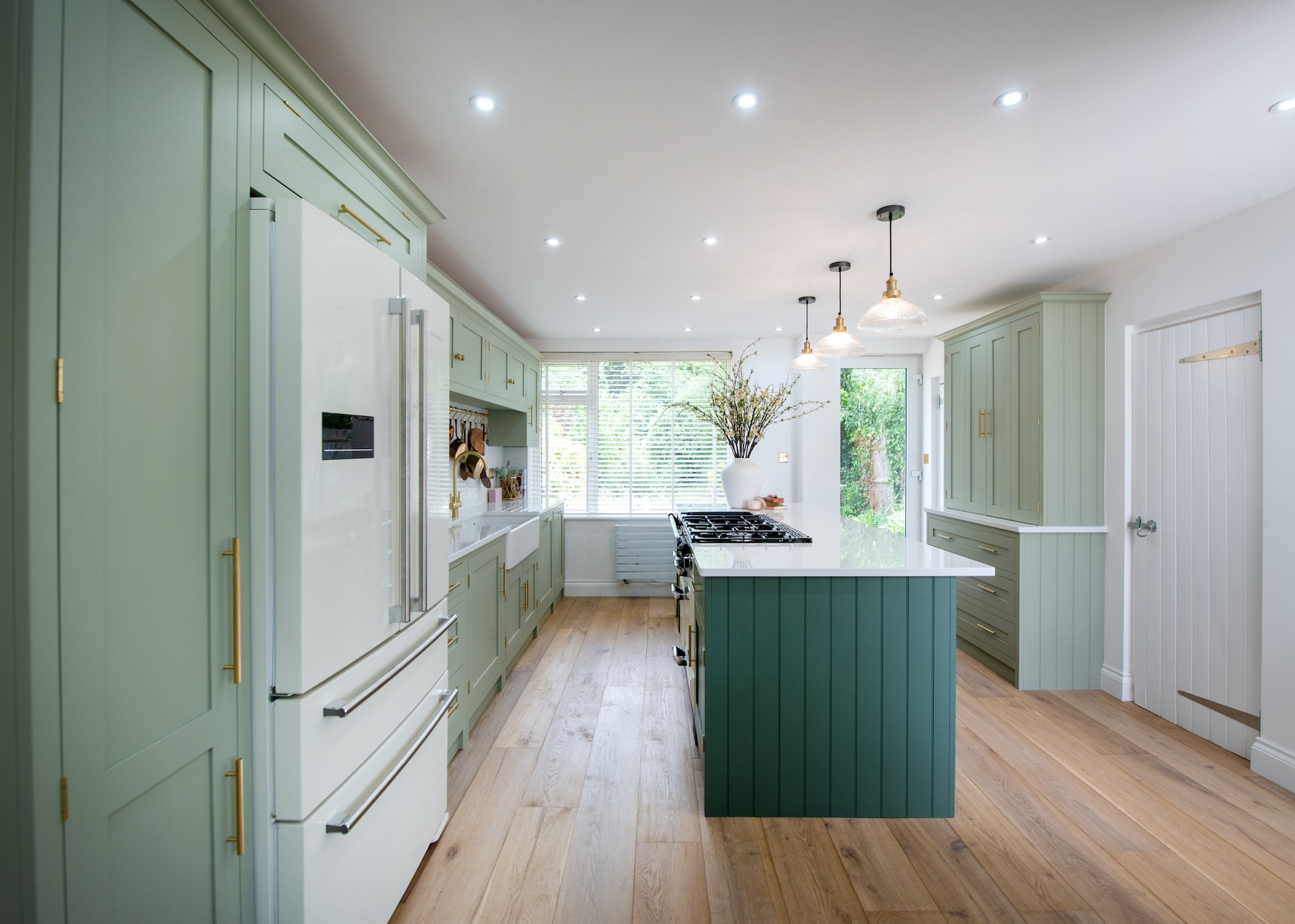 A light green kitchen with an island int he middle that has a hob on top. Above the island are pendant lights. You can also see a double door fridge on the left side of the kitchen.