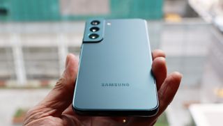 En hand håller upp en blågrå Samsung Galaxy S22 med baksidan vänd mot kameran.