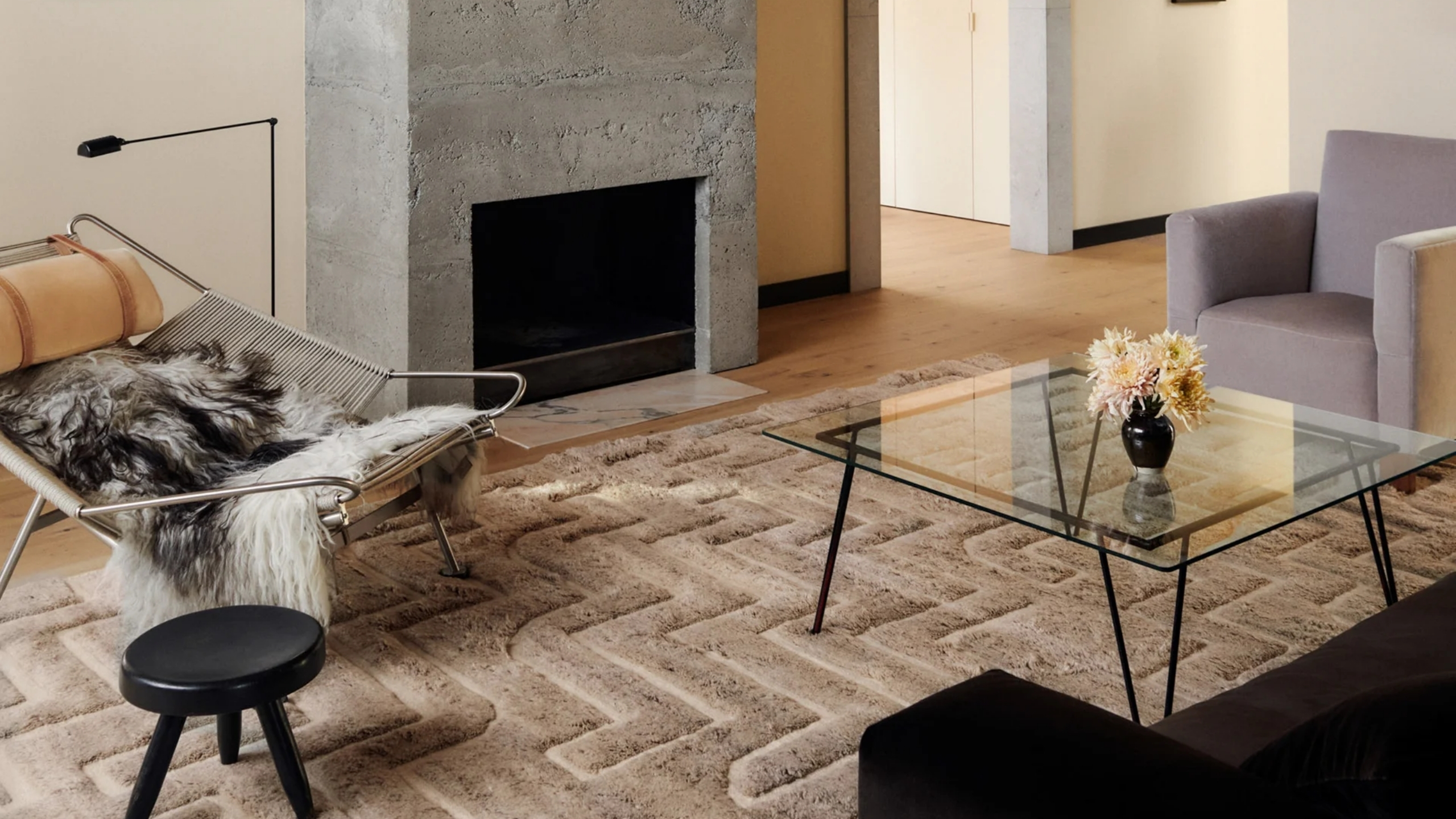 Neutral living room with a sculptural embossed rug, glass coffee table, lounge chair, concrete fireplace and soft upholstered seating
