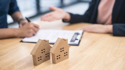 A pair of model homes sit on a conference room table while two people sign documents, only their hands showing.