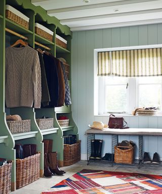 Boot room with blue paneled walls, painted green bespoke open storage unit with coat hooks, seating, wicker baskets, stone flooring, red patterned rug, window with cream and green patterned fabric blind