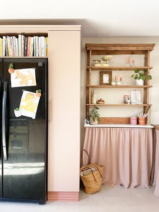 A light pink kitchen with a black fridge with a built-in shelf surround and wooden shelves on the wall