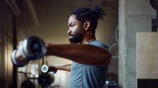 A man performs a lateral raise with dumbbells in a gym. He is standing with a dumbbell in each hand, holding them out to the sides. 