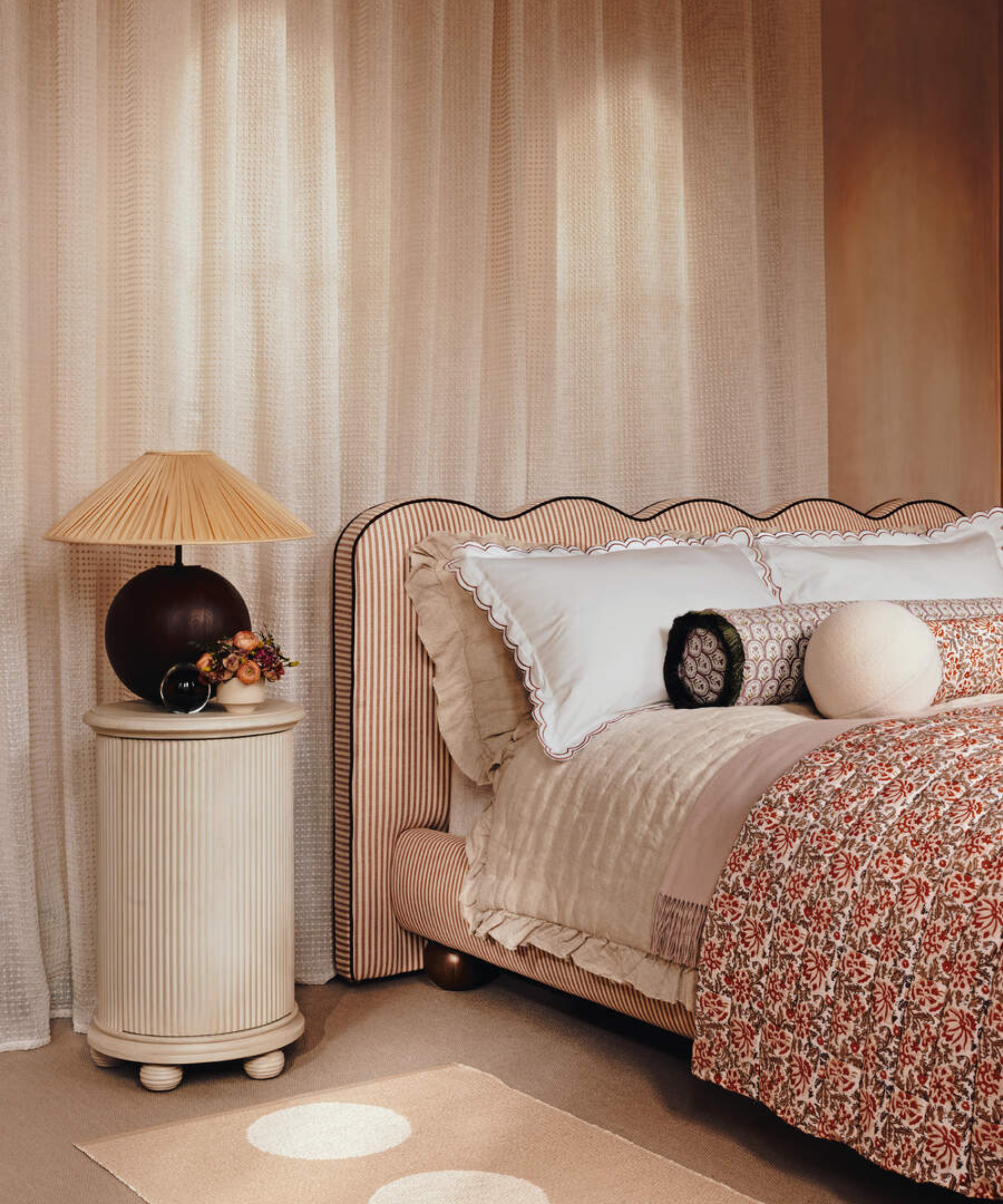 Blush pink bedroom featuring a striped fabric upholstered bed frame with a wavy headboard, patterned bedding, a fluted nightstand topped with pleated shade table lamp, and curtains