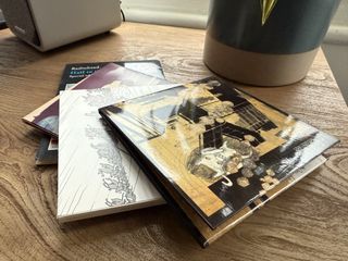 A pile of CDs on a desk. A speaker and plant can be seen in the background.