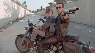 The Terminator, Arnold Schwarzenegger, looks back and points a single-barrel shotgun while on a motor cycle