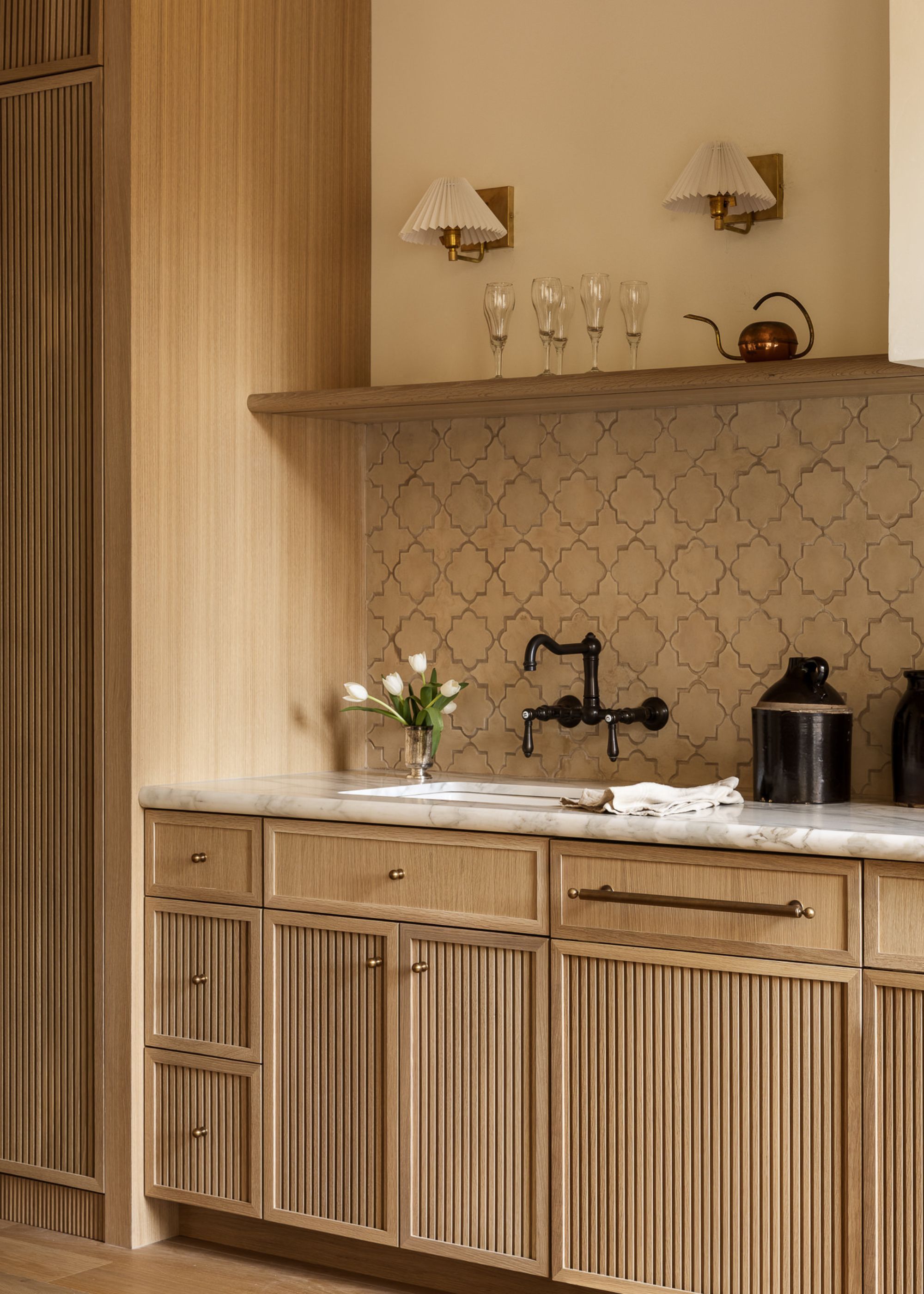 A modern timber kitchen with fluted detailing on the cabinetry, a marble worktop, a black wall-hung tap and a display shelf below two small brass-based wall lights with white pleated shades