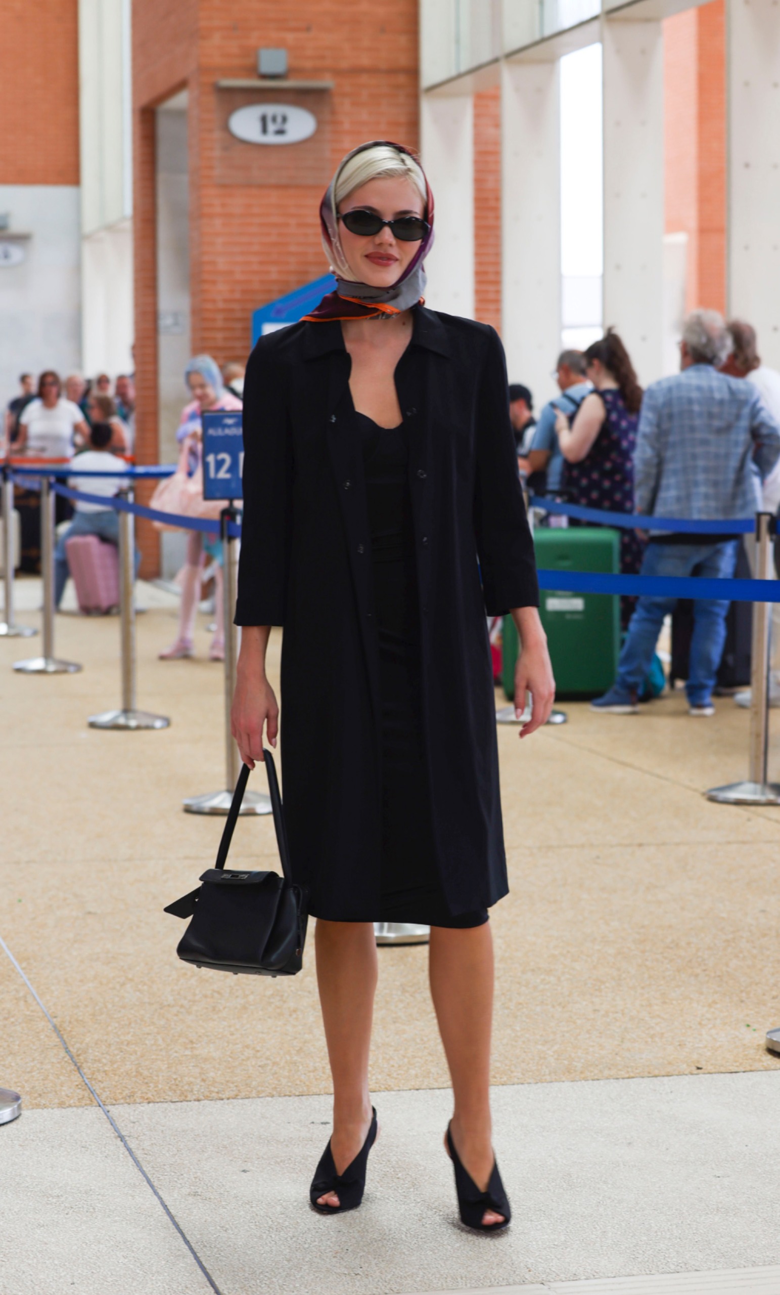 An image of Hayes Warner wearing an LBD with peep-toe shoes, oval sunglasses and a scarf whilst at the Venice Airport.