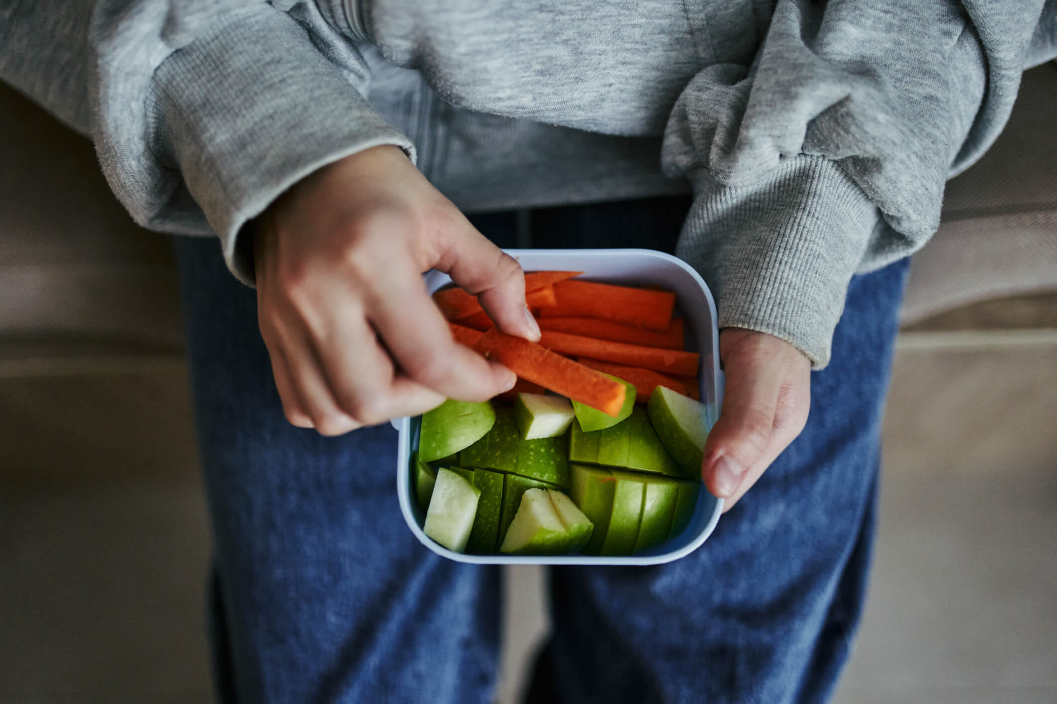 A child holding a small Tupperware box containing carrot sticks and green apple chunks.