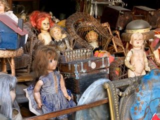 Messy packed room full of antique objects like dolls, an accordion, wicker or basket chairs 