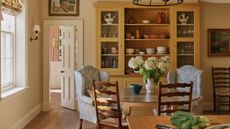Farmhouse kitchen with wooden floors, a yellow dresser filled with ceramics, a round wooden dining table with two armchairs and dining chairs, and a wooden island with a bowl of fruit on top.