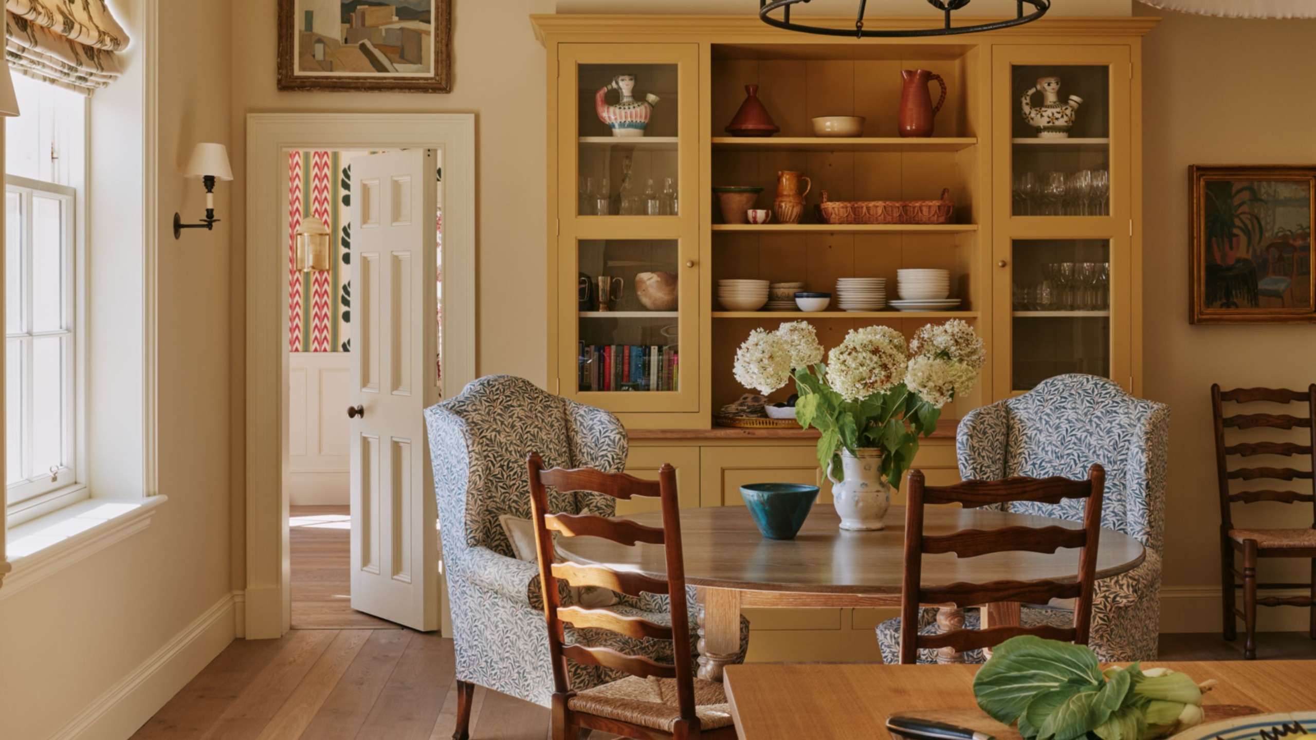 Farmhouse kitchen with wooden floors, a yellow dresser filled with ceramics, a round wooden dining table with two armchairs and dining chairs, and a wooden island with a bowl of fruit on top.