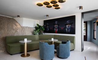 Interior view of the seating area at Bar Roc at Ellerman House featuring white walls, green plants, multiple ceiling lights, a green corner sofa, two curved blue chairs, grey and bronze coloured round tables and art on the wall of three male characters in hats and suits