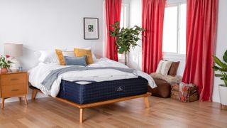 The DreamCloud Mattress shown on a light wooden bed frame in a bedroom with red curtains