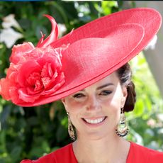 Kate Middleton wearing a red hat with flowers and dangling earrings smiling in front of flowers