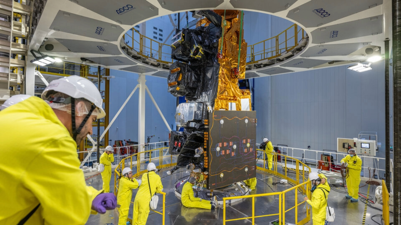 A bunch of people wearing bright yellow jumpsuits and white hard hats stand around a large vertical golden covered rocket. 
