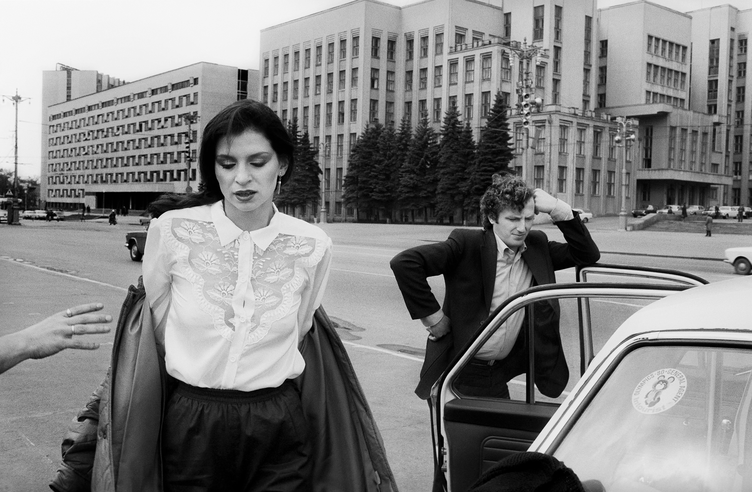 Black and white photo of a woman in a lace blouse and man in a suit near a car on a city street with large buildings in the background