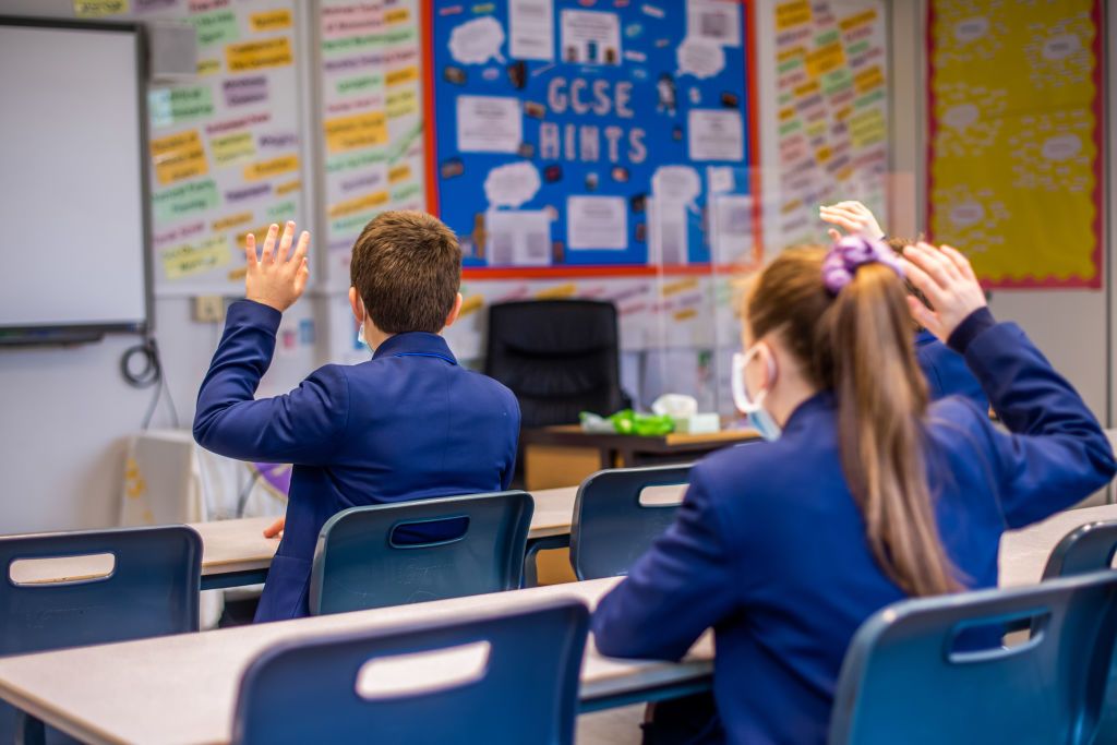 Children at school after lockdown ends, in the classroom with face masks on and hands raised