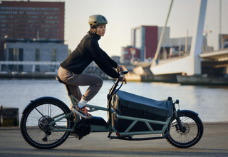 A woman dressed in black rides a front loading Riese and Muller cargo bike along a dock front.