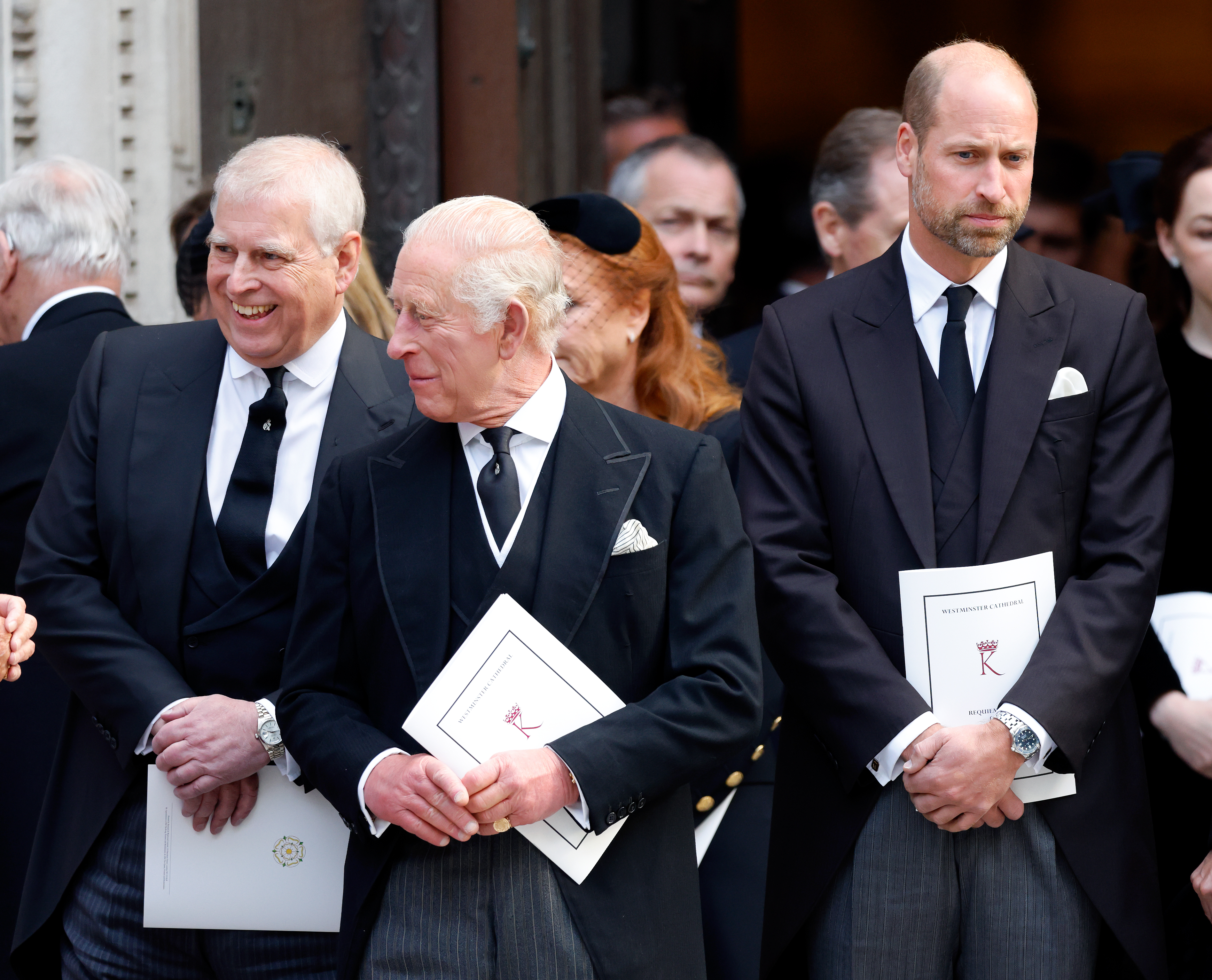 Prince Andrew and King Charles laughing and holding programs standing next to Prince William, who is looking away