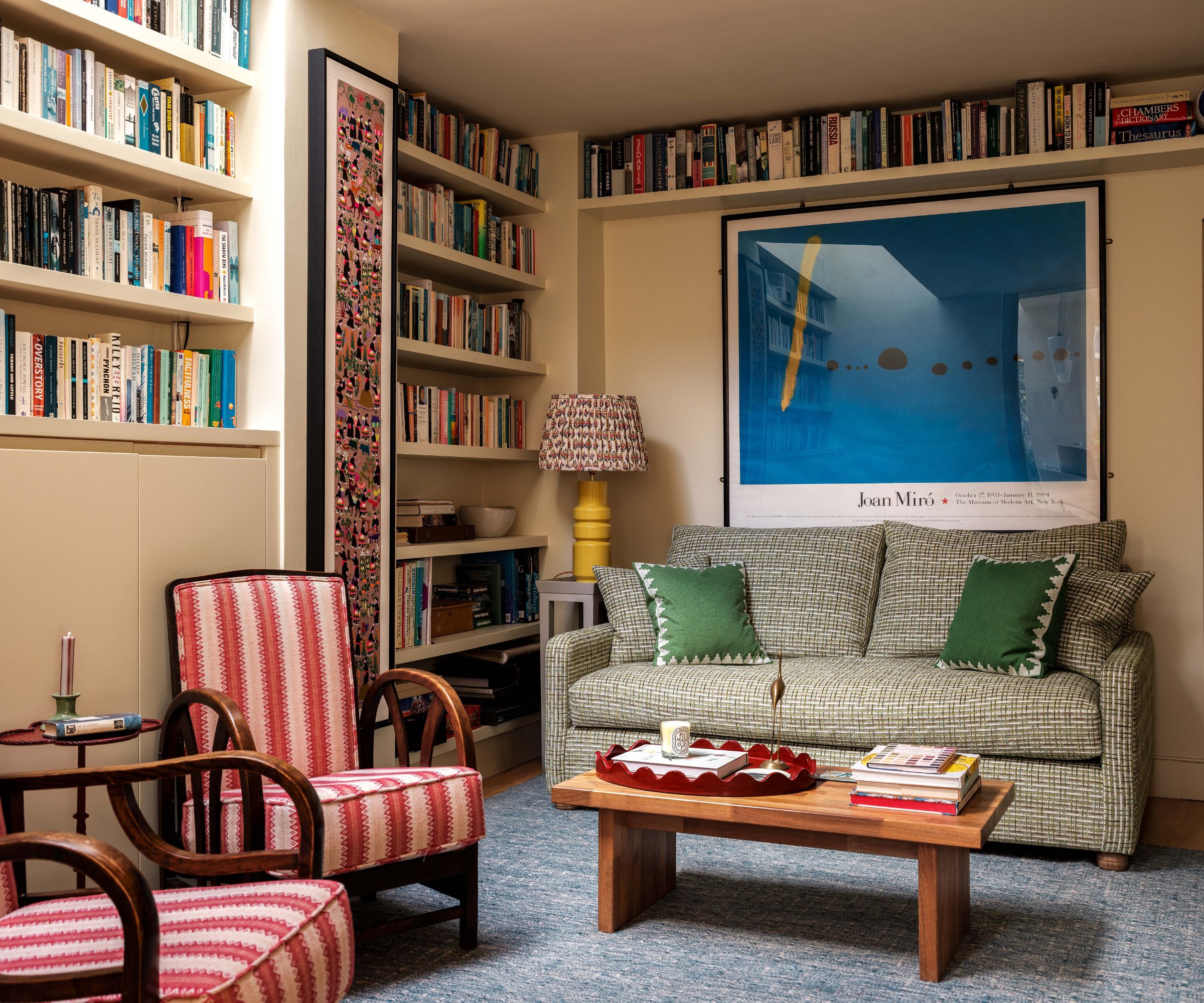 Living room with pale beige walls, built-in bookshelves and an alcove of books, a Joan Miro print above a green sofa, a wooden coffee table and two pink striped armchairs.