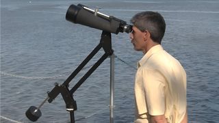 A man stands in front of a body of water looking through a pair of binoculars mounted on a tripod with a counterweight.