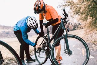 This image shows two people removing a rear wheel from a bike one is wearing an orange helmet and jersey, the other is in a blue jersey with a white helmet. They are on a road with an autumnal background of trees