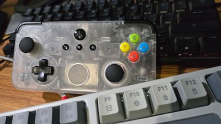 A Classic Clear CRKD NEO S rectangular controller sits on a desk between one black keyboard and one grey keyboard. The controller takes up much of the frame, but is clearly much smaller than both of the keyboards. The controller body is made of cloudy, clear plastic, allowing the viewer to see the internals.