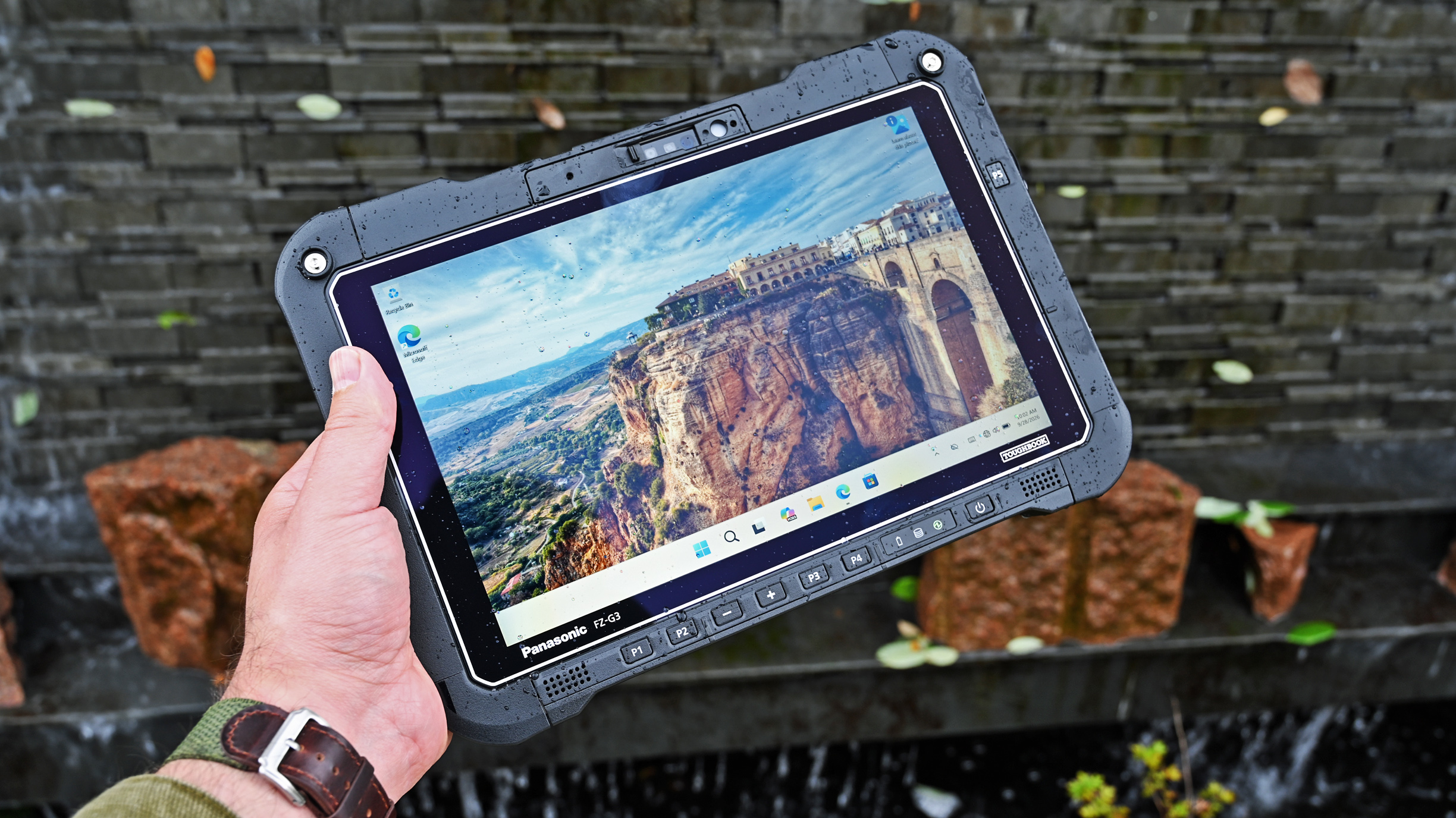 A hand holding a water-splattered Panasonic TOUGHBOOK G3 tablet outdoors, featuring a bright Windows screen with landscape wallpaper.