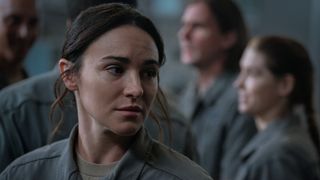 Close up of a worried-looking woman with long dark hair tied up. She is wearing a gray jumpsuit. In the background you can see several other people also wearing the same gray jumpsuit.