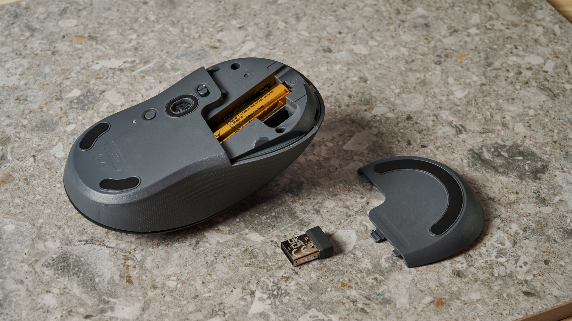 the underside of a black mouse with a scroll wheel, two side buttons, and grooved edges, with a logi logo, sits atop a pale table with a blue background