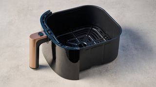 the cooking basket of a large black 5.3 quart capacity air fryer with black touchscreen and rose gold embellishments sits upon a brown table with a blue background